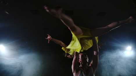 Two-dancers-a-man-and-a-woman-run-to-each-other-and-a-male-partner-raises-a-woman-in-a-yellow-dress-in-her-arms-and-rotates-in-the-air-performing-top-support.